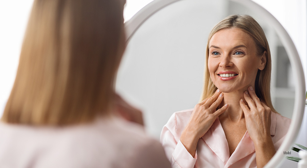 woman touching her neck with both hands as she looks at her reflection in the mirror.